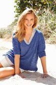 A young woman wearing shorts and a blue mottled shirt sitting on a beach