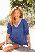 A young woman wearing shorts and a blue mottled shirt sitting on a beach