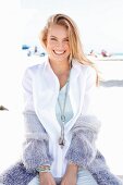 A blonde woman on a beach wearing a white blouse and a purple cardigan