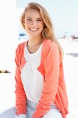 A blonde woman on a beach wearing a white sweater and a salmon coloured jacket