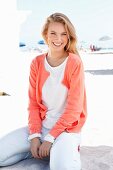 A blonde woman on a beach wearing a white sweater and a salmon coloured jacket