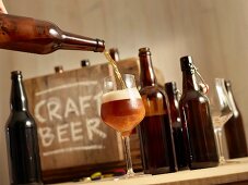 Bier wird eingeschenkt, im Hintergrund geöffnete Bierflaschen Holzkiste mit Aufschrift 'Craft Beer'