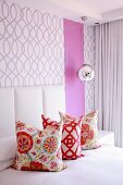 Three colourful scatter cushions on bed against wall covered in purple-patterned wallpaper