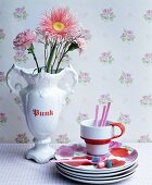 Tellerstapel mit Rosenmuster und Tasse neben Blumenstrauss in weisser Henkelvase vor nostalgischer Rosentapete