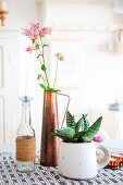 Rosa Akeleiblüten in Kupfervase, Glasflasche mit Kerze und Sukkulente in rustikaler Tasse