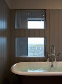 Freistehende Badewanne mit Wasser vor Holzverkleidung und Innenfenster
