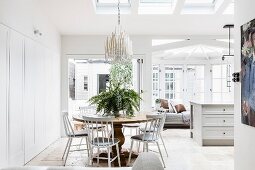 Dining area with white chairs around a round wooden table in an open living room, in the background a kitchen counter and lounge