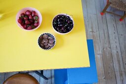 Three bowls of strawberries, cherries and chocolate biscuits on yellow table above blue step-stool