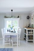 White dining table and chairs next to shelves of crockery below window in Swedish house