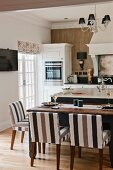 Island counter, set dining table and chairs with striped brown upholstery in white country-house kitchen