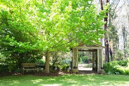 Idyllischer Gartenplatz mit Holzpavillion und Gartenbank unter grünem Ahornbaum