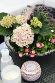 Pflanzschale mit Hortensien und Bellis neben weissen Kerzen und dekorativem Tiegel