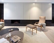 Set of coffee tables, armchair with wooden frame and matching footstool in front of white fitted cupboards with integrated gas fireplace