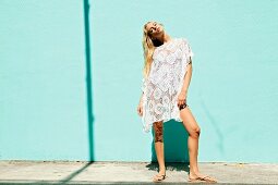Junge blonde Frau mit Spitzen-Tunika über Bikini an türkiser Wand
