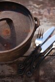 A rustic cast-iron saucepan and cutlery on a wooden table