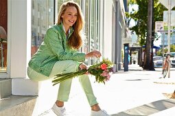 A young blonde woman sitting on a step outside a shop window holding a bunch of flowers