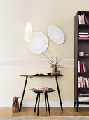 Elegante Wanduntergliederung mit Stuckleisten und pastellfarbenen Vliestapeten, davor schwarzer Tisch mit Hocker und Bücherregal
