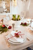 White place setting with silver cutlery and red cord around pine cone on festively decorated dining table