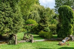 Wooden deckchair in green garden with various trees, pond and rustic bridge