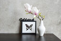 Magnolia flowers in white vase in front of picture of butterfly leaning against concrete wall