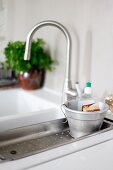 Washing-up liquid in metal pot with hooks over edge on draining rack next to sink