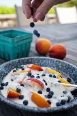 A hand sprinkling blueberries onto yoghurt with berries and peach slices