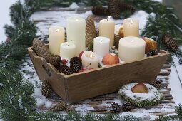 Holzkiste mit Kerzen, Zapfen, Äpfeln (Malus) und Moos im Schnee