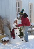 Frau baut Schneemann mit Tontopf als Hut, Schal, Kohlstrunk als Pfeife