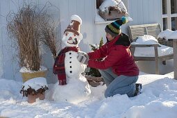 Frau baut Schneemann mit Tontopf als Hut, Schal, Kohlstrunk als Pfeife