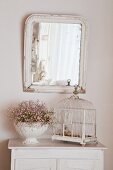 Vintage birdcage an urn of flowers on white cabinet below mirror on wall