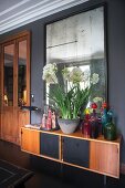 Colourful glass bottles and bowl of white-flowering plants on retro sideboard below mirror on wall