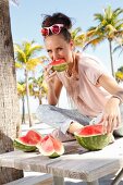 Brünette Frau in cremefarbenem T-Shirt und hellgrauer Hose isst am Strand eine Melone