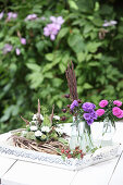 Wreath decorated with white asters, thistles and poppy seedheads next to pink asters in glass bottles