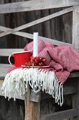 Candlestick decorated with rose hips next to mug arranged on fringed blanket and rustic wooden bench