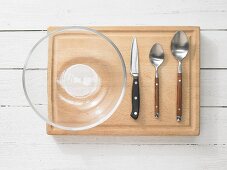 Kitchen utensils: glass bowl, two spoons, kitchen knife and wooden chopping board