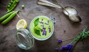 Green gazpacho in jar with peas and lavender on wooden background with vintage spoons