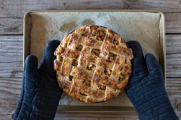 A freshly baked (still warm) caramel apple pie is placed on the table by a person wearing oven mitts