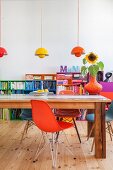 Shell chairs of various colours at long wooden table in front of colourful shelves