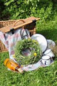 Picnic basket, blanket, enamel crockery and wreath of wildflowers on green lawn