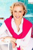 A blonde woman wearing a white blouse and a pink jumper draped over her shoulders next to a pool