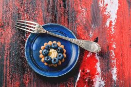 Top view on blueberry mini tart served on blue ceramic plate with vintage fork over black and red wooden background