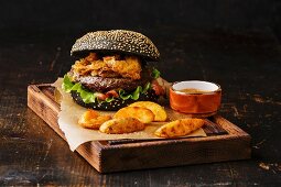 Black burger with sesame seed bun, meat, bacon, onion fries rings and potato wedges on dark wooden background