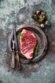 Rohes Striploin Steak mit Rosmarin und Vintage-Besteck auf Teller
