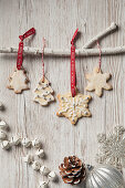 Edible Christmas tree decoration biscuits in the shape of trees and snowflakes iced and haning by red Noel ribbon and bakers twine from a white branch on a rustic white wood background and other Christmas decorations