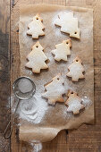 Tree shaped biscuits on baking paper being dusted with icing sugar from a mini sifter on a rustic wooden surface
