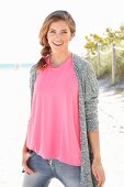 Blonde Frau in pinkfarbenem Top, grauer Strickjacke und Jeanshose am Strand