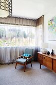 Upholstered armchair and fifties style sideboard in front of window with airy curtain