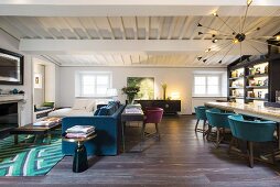 Blaues Polstersofa vor Kamin und langer Tisch mit Schalenstühlen in offenem Wohnraum mit weisser Holzbalkendecke