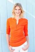 A blonde woman wearing an orange long-sleeved top with zip and white trousers