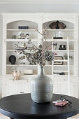 Dried flowers in stoneware vase on round table in front of shelves